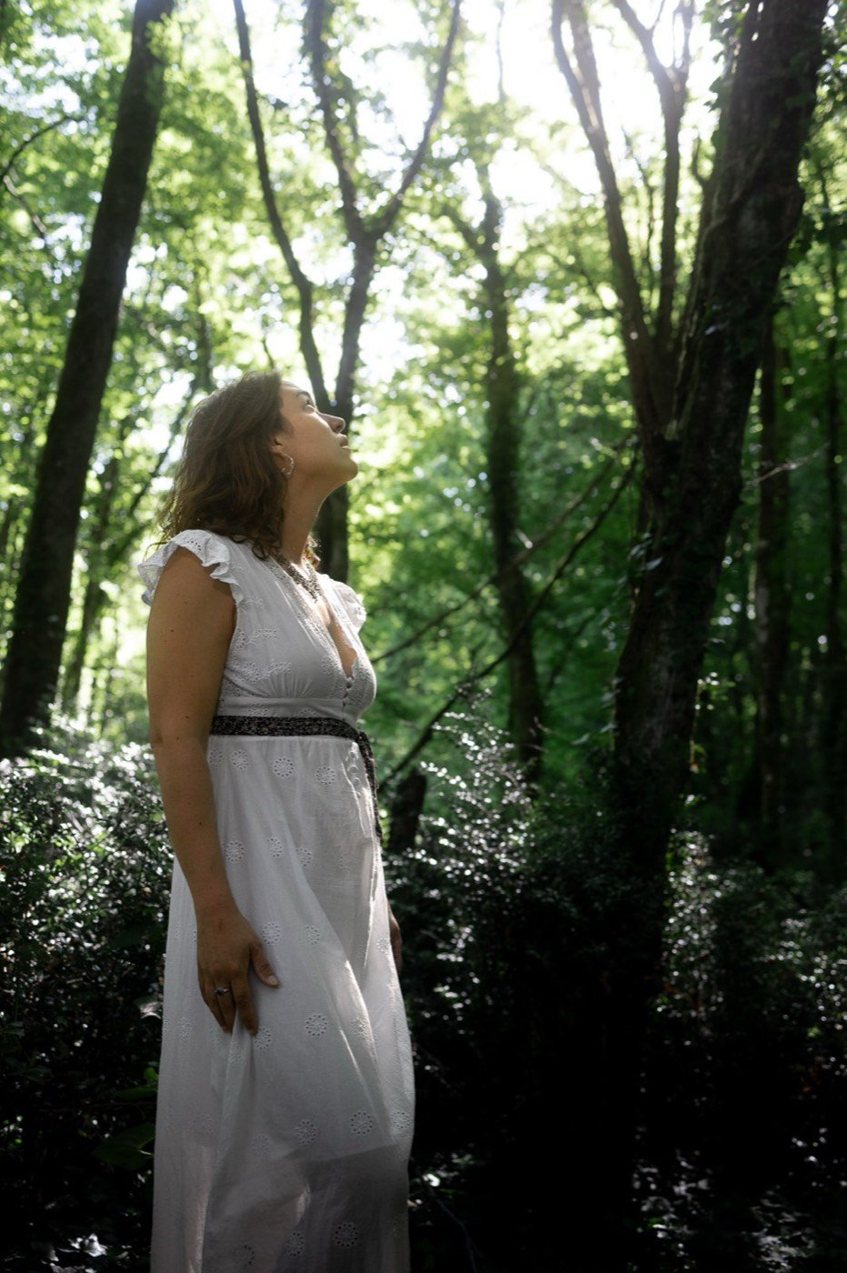 Jade en robe blanche, le regard levé vers la lumière qui traverse les arbres
