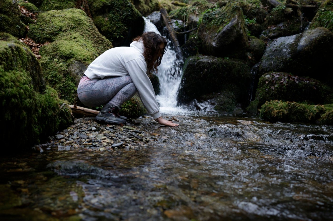 Jade accroupie au bord d'une cascade, la main dans l'eau d'une rivière moussue